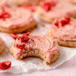 A strawberry cookie topped with strawberry buttercream and crumbled freeze-dried strawberries with a bite removed.