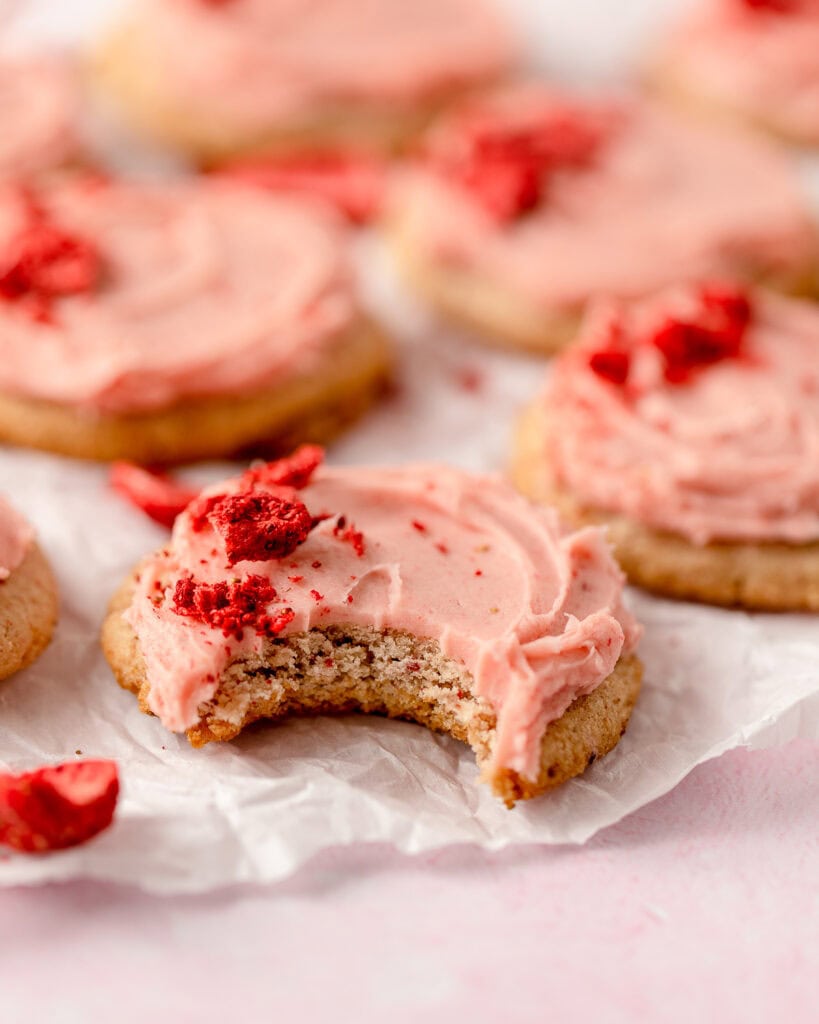 A strawberry cookie topped with strawberry buttercream and crumbled freeze-dried strawberries with a bite removed.