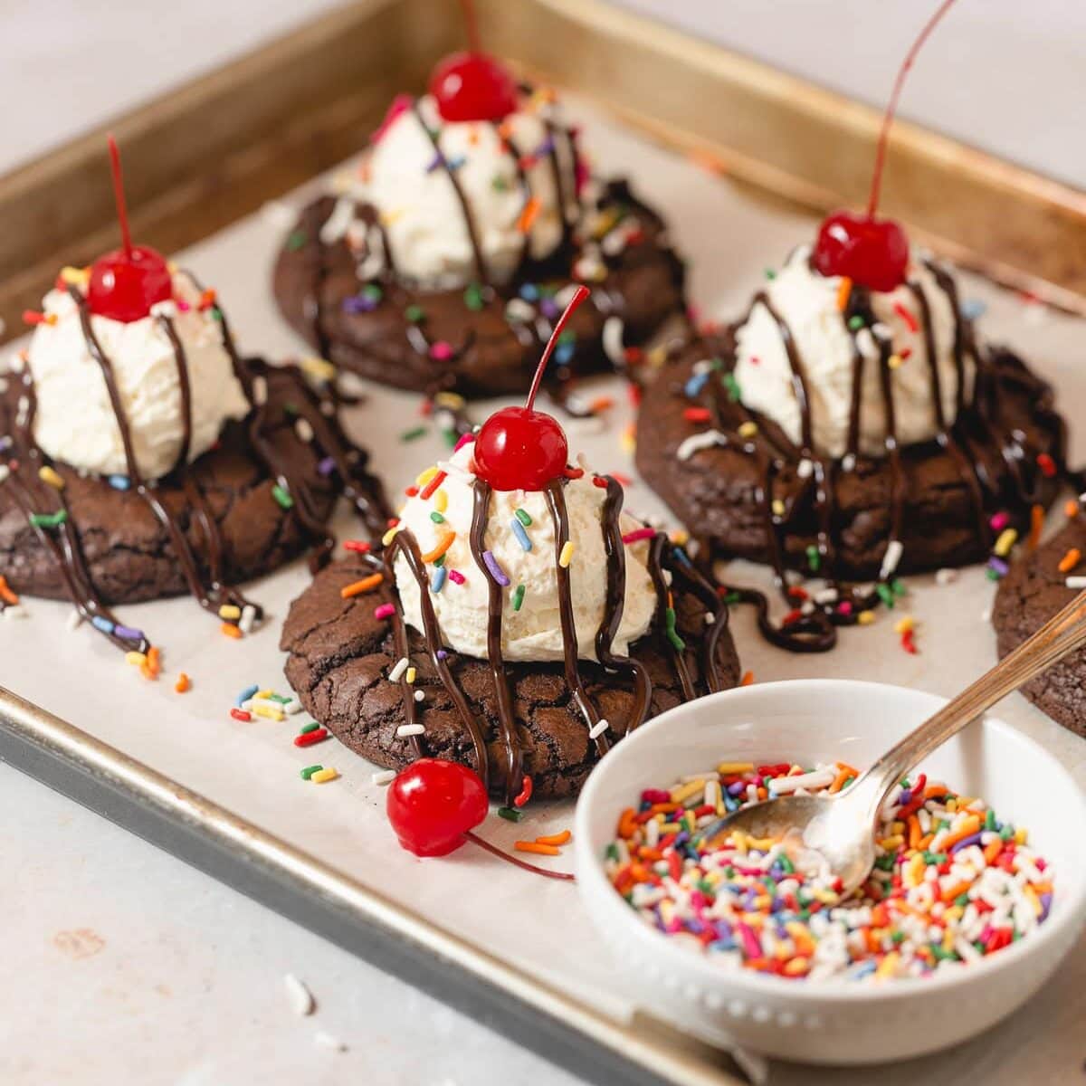 Brownie cookies topped with vanilla mousse, hot fudge drizzles, sprinkles, and cherries on top.