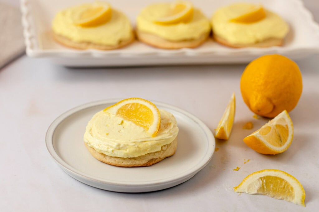 A lemonade sugar cookie topped with yellow lemon buttercream and a lemon slice with more lemons surrounding.