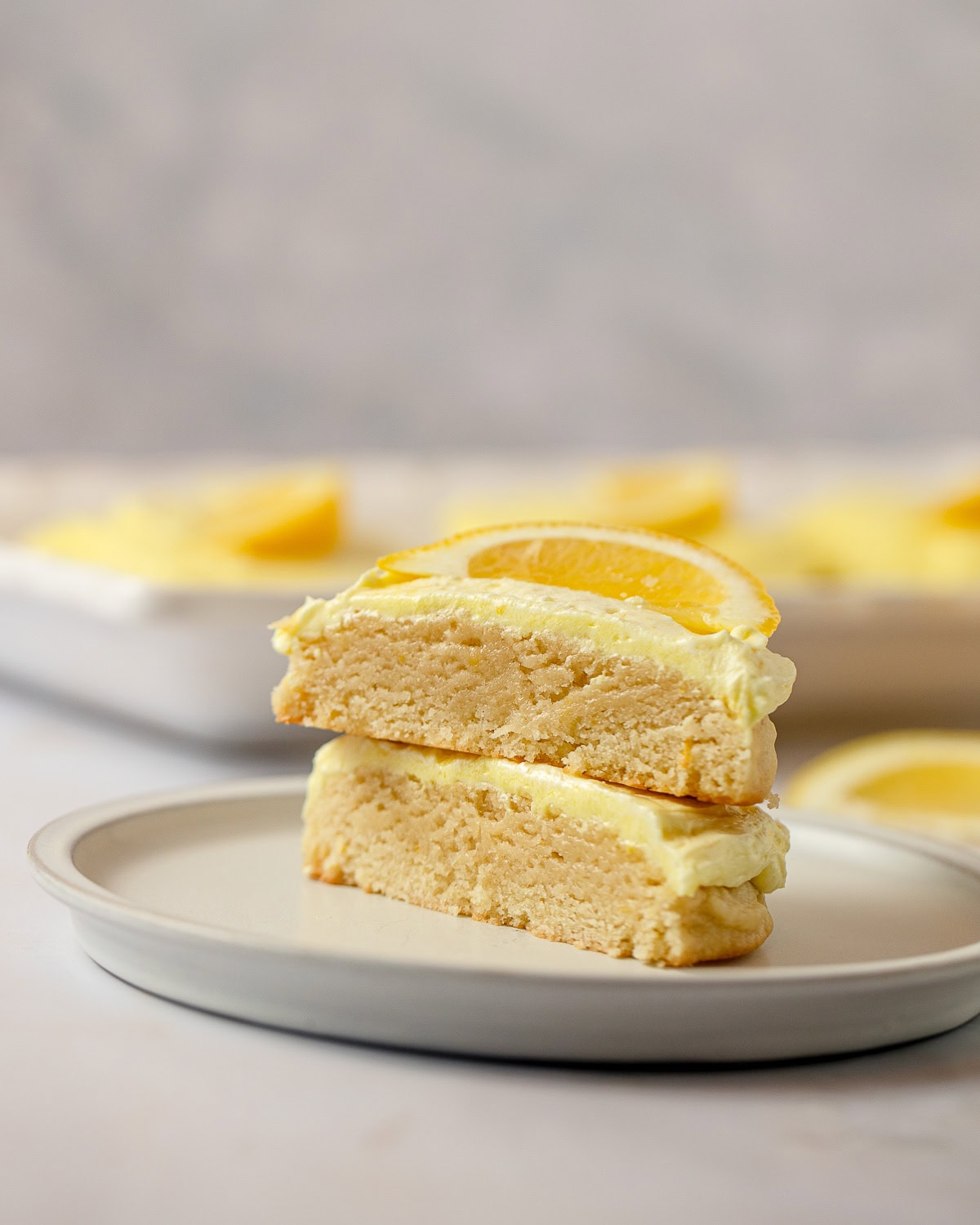 A lemon cookie with yellow frosting and a lemon slice on top cut in half with the halves stacked on top of each other.