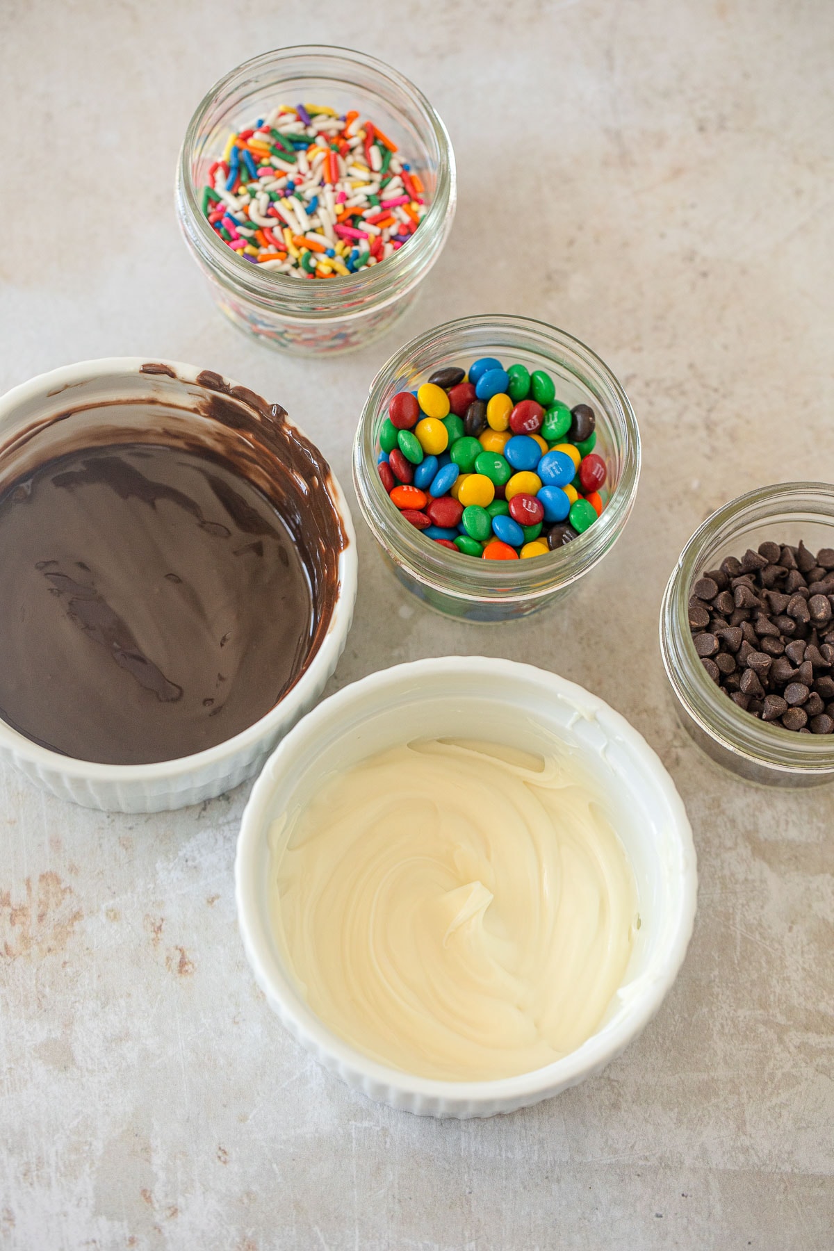 Bowls of: melted white chocolate, melted chocolate chips, mini chocolate chips, mini m&ms, and sprinkles.