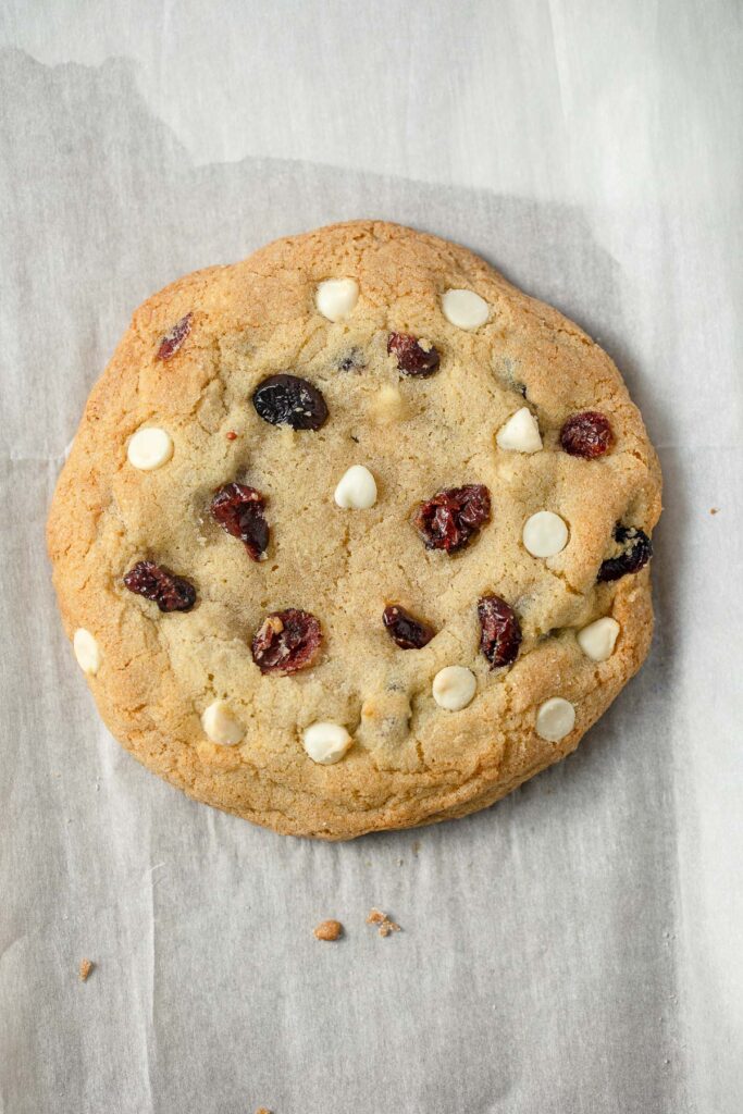 A cookie with dried cranberries and white chips on a parchment-lined baking sheet.