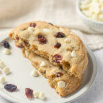 A cookie with dried cranberries and white chips broken in half with the halves stacked on a plate.