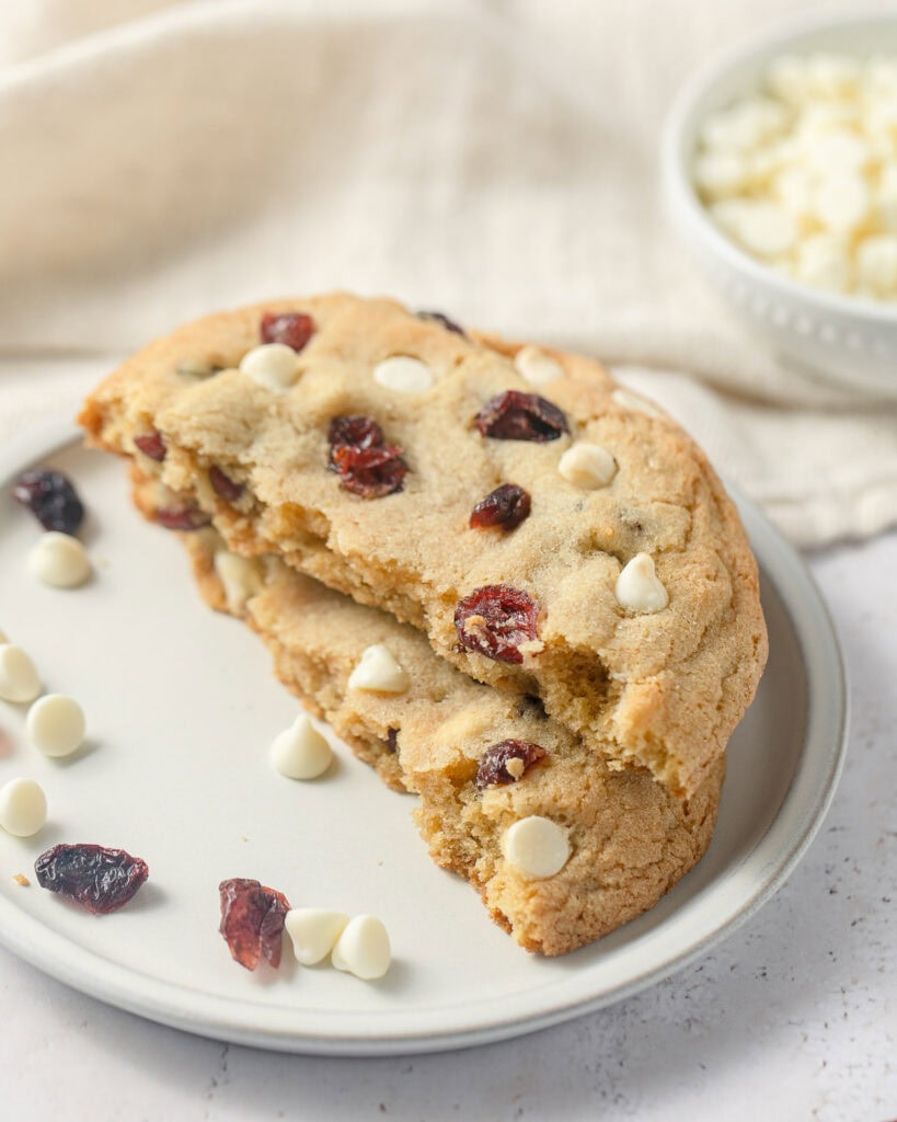 A cookie with dried cranberries and white chips broken in half with the halves stacked on a plate.