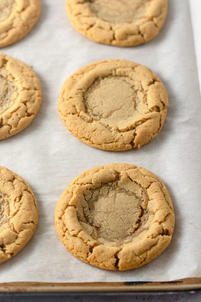 A baking sheet lined with parchment with peanut butter cookies that have a divot pressed into the middle for filling.