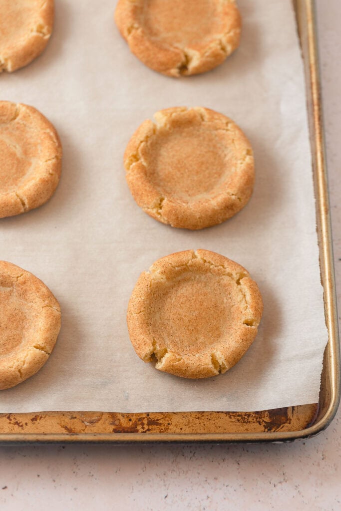 Snickerdoodle cookies with large divots pressed into the middle on a parchment-lined baking sheet.