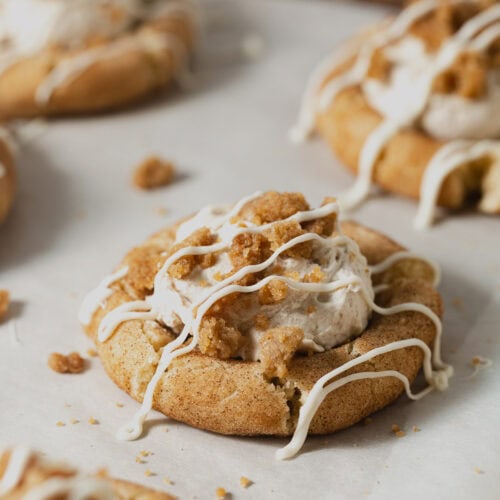 Snickerdoodle cookies filled with cinnamon whipped cream and topped with streusel and drizzles of white chocolate on a parchment-lined baking sheet.