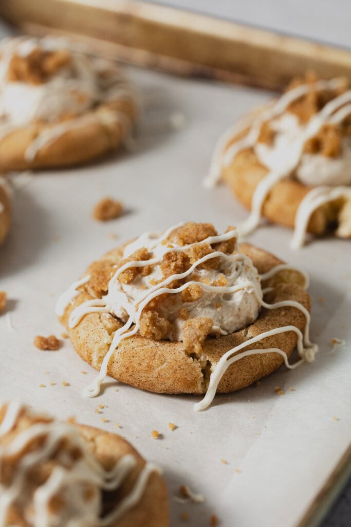 Snickerdoodle cookies filled with cinnamon whipped cream and topped with streusel and drizzles of white chocolate on a parchment-lined baking sheet.