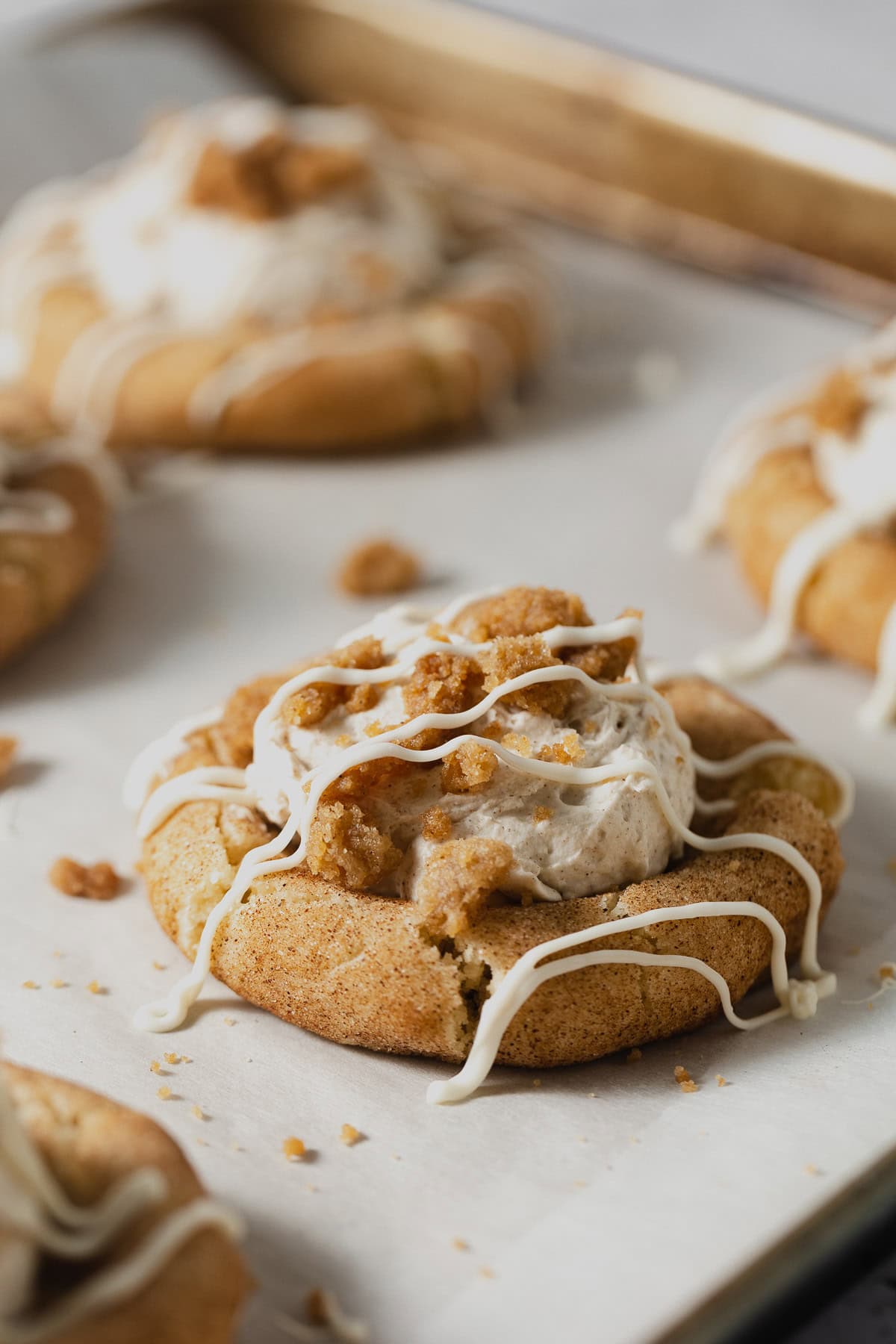 Snickerdoodle cookies filled with cinnamon whipped cream and topped with streusel and drizzles of white chocolate on a parchment-lined baking sheet.