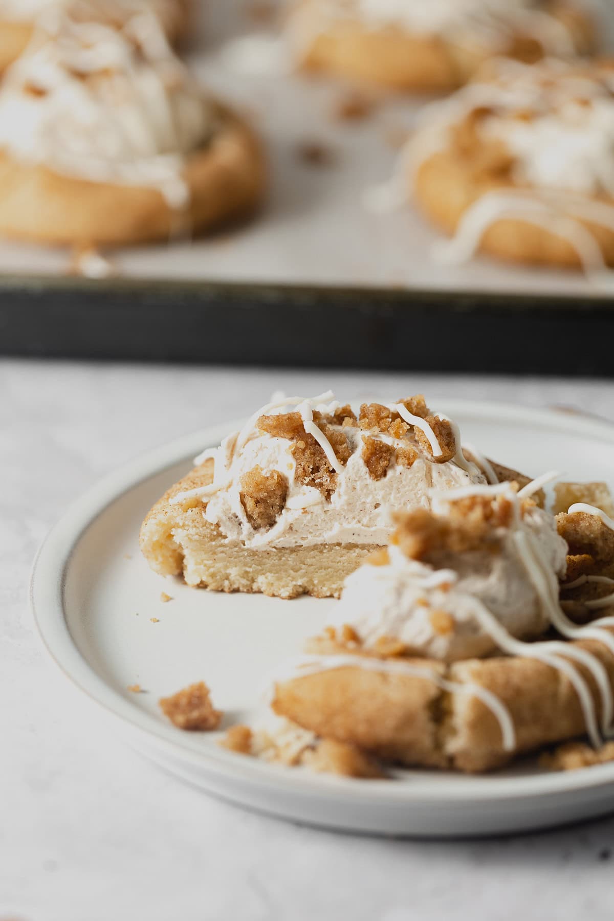 Halves of a snickerdoodle cookie filled with cinnamon whipped cream and topped with streusel and drizzles of white chocolate and served on a rustic plate.