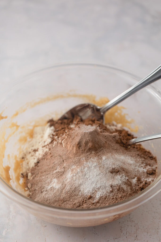 A bowl of cookie dough with the dry ingredients (flour, hot chocolate mix, natural cocoa powder, baking soda, salt) added on top.