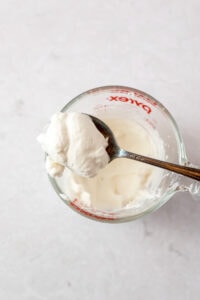 A spoon filled with whipped cream resting on top of a glass measuring cup filled with more whipped cream.