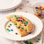 A single M&M cookie split in half and stacked on a white plate with more mini M&Ms surrounding.