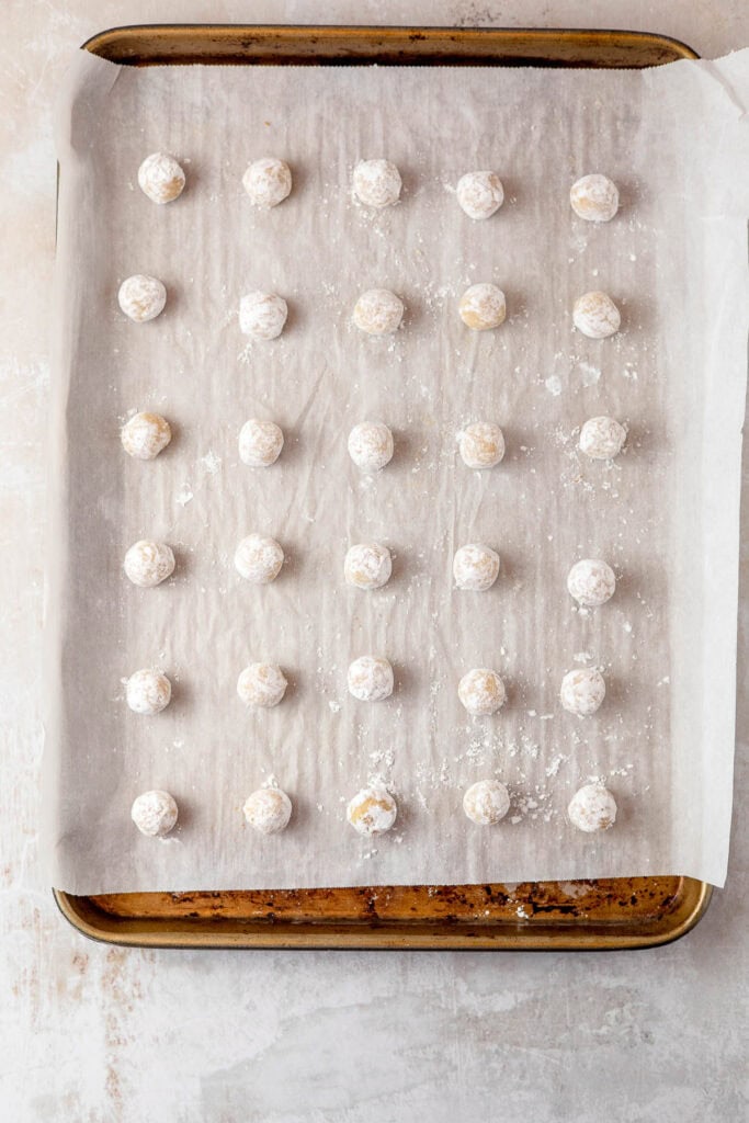 A parchment-lined baking tray of cookie dough scoops rolled in powdered sugar.