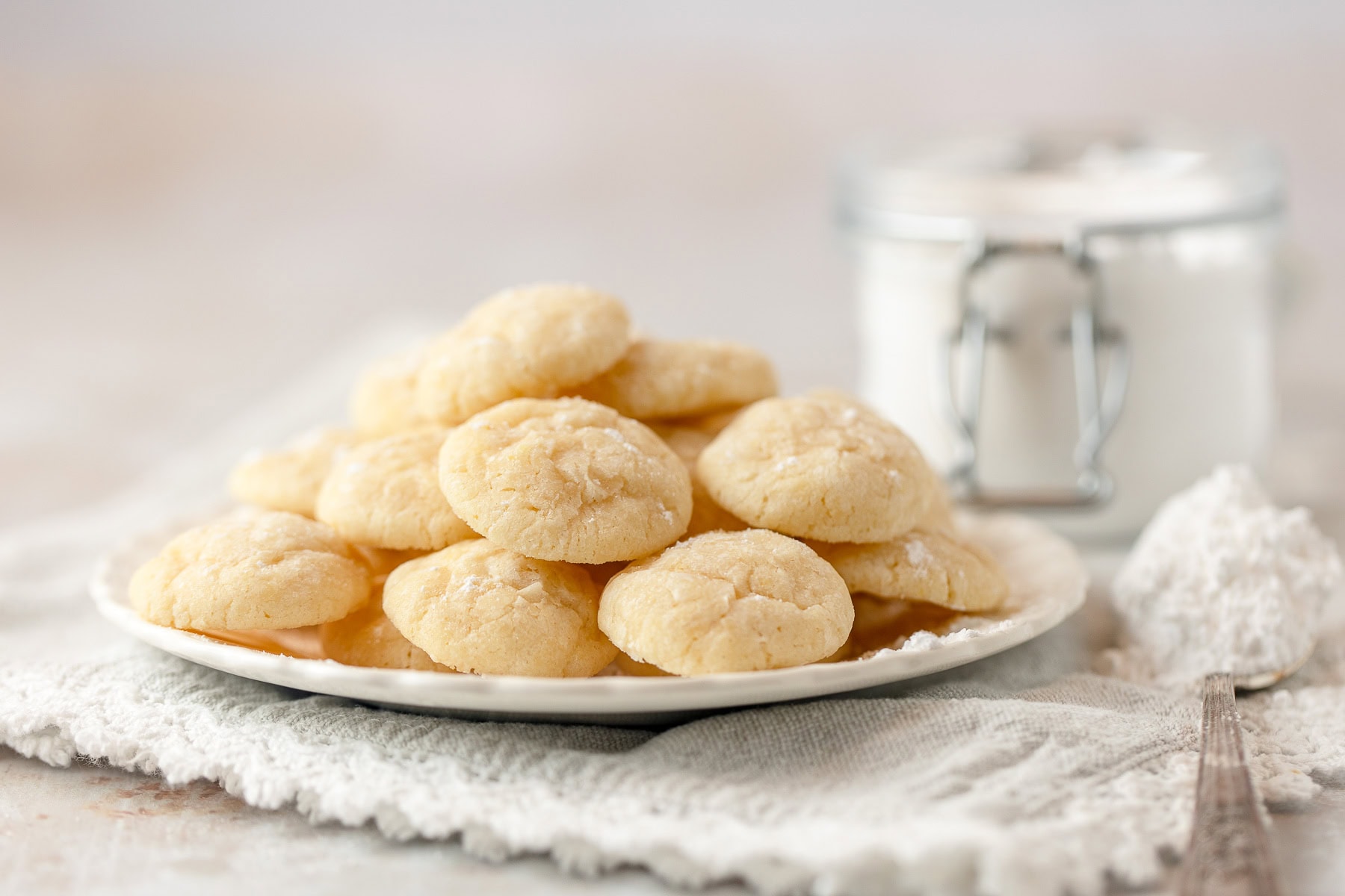 A plate filled with mini lemon cookies surrounded with a jar of powdered sugar and a spoonful of powdered sugar.